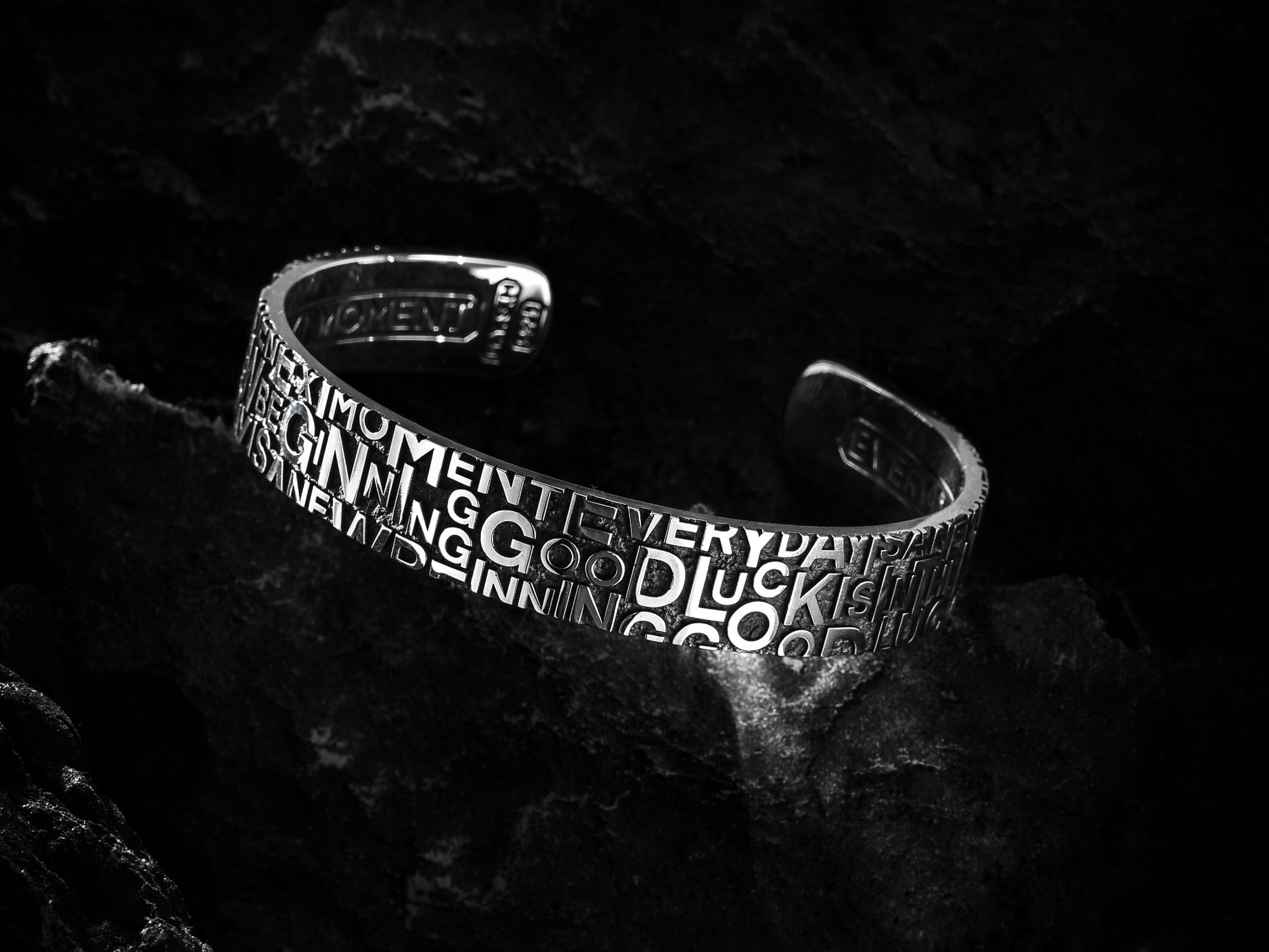 A silver cuff engraved with overlapping letters, resting on black rock